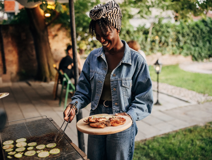 Woman barbequing vegetables and chicken in her backyard with people seated at a table in the background